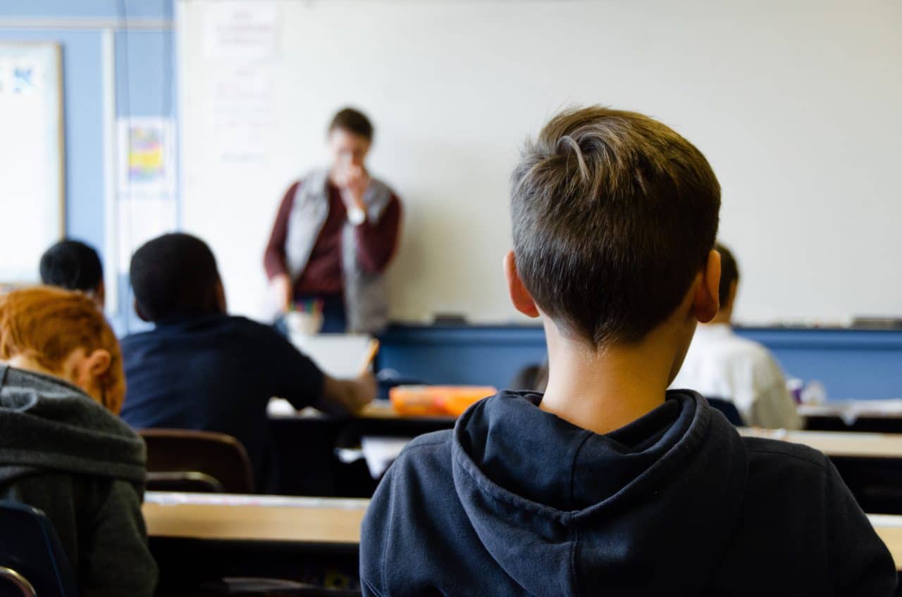 Children learning together in a classroom while a teacher stands up front.