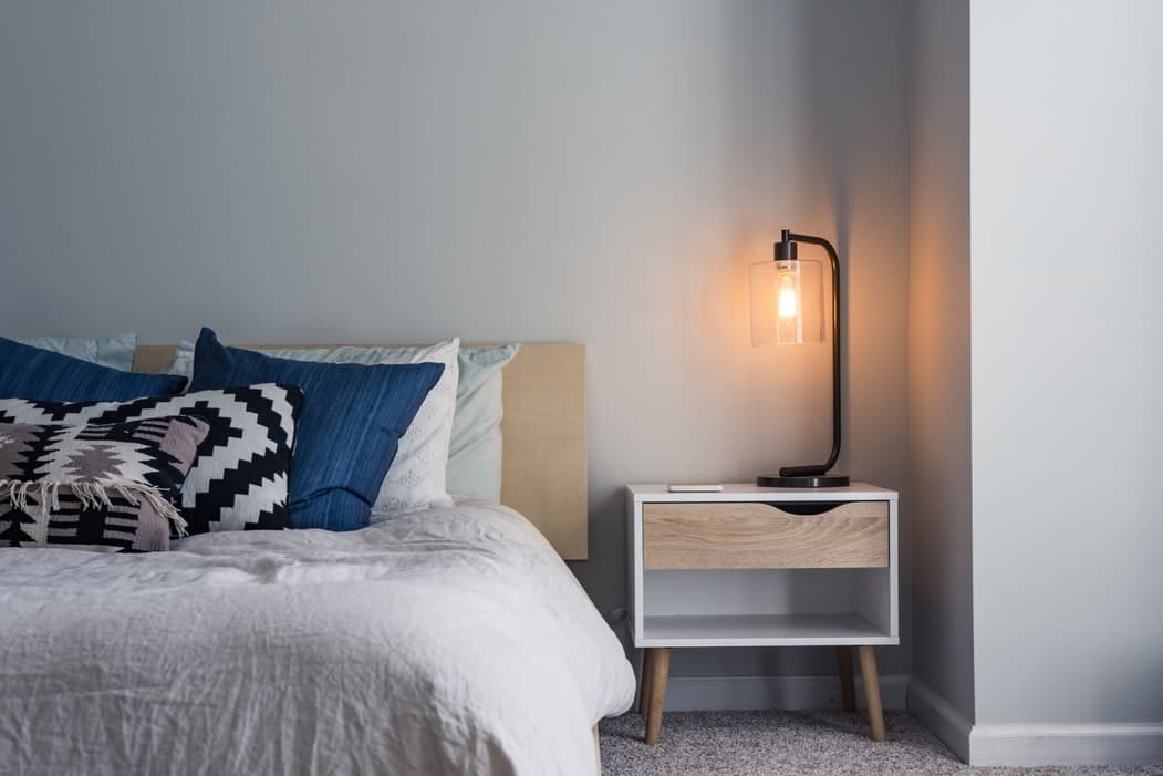 A picture of a night stand with a lamp next to a bed.