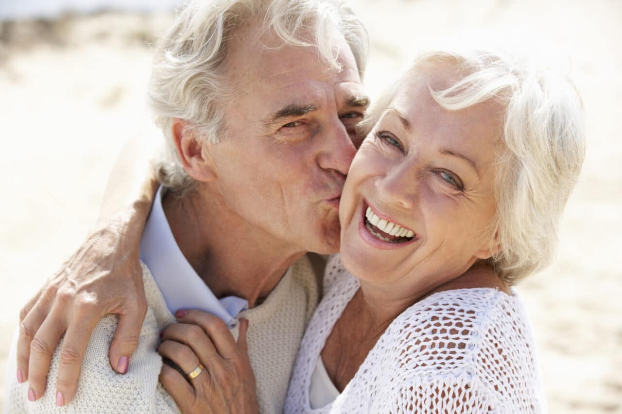 Senior Couple Walking Along Beach Together Smiling To Camera.